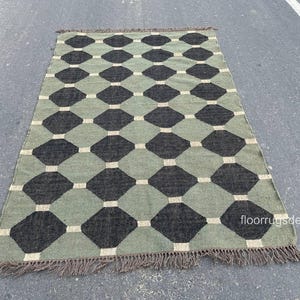May include: A rectangular rug with a geometric pattern of black and green diamonds connected by white squares. The rug has a fringe on the short ends and is made of a woven material. The rug is on a gray surface.