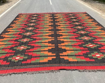 May include: A rectangular area rug featuring a geometric pattern with repeating diamond shapes in black, red, orange, and olive green. The rug has a fringed edge on the bottom and a symmetrical design with a central vertical black stripe.