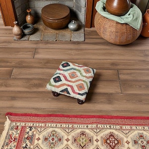 May include: A small, square footstool with a geometric pattern in shades of red, orange, green, and blue. The footstool has dark wooden legs and is placed on a wooden floor. The image also shows a decorative rug.