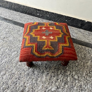 Könnte beinhalten: Ein quadratischer, niedriger Hocker mit gemusterter Oberseite. Das Design zeigt eine zentrale Kreuzform und geometrische Muster in Rot-, Braun-, Gelb- und Weißtönen. Der Hocker wird von vier kurzen, dunklen Holzbeinen getragen.