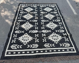 May include: A rectangular black rug with a white geometric pattern. The rug features a repeating design of diamond shapes, arrows, and floral motifs. The edges are finished with a fringe.