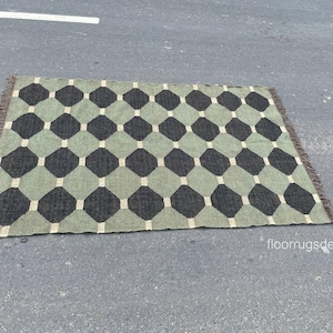 Puede incluir: Una alfombra rectangular con un patrón geométrico de diamantes negros y octágonos verde claro. La alfombra tiene un borde con flecos y se muestra sobre una superficie gris. El diseño de la alfombra se repite.