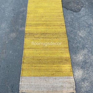 May include: A rectangular yellow and beige woven rug. The rug has a yellow center with a beige border at each end. The text "floorrugsdecor" is at the bottom of the rug.