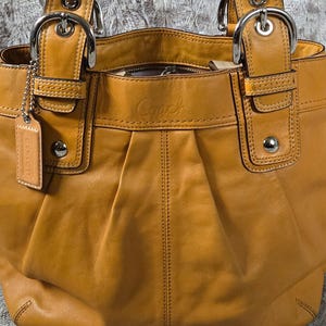 May include: A mustard-colored leather handbag with two top handles and silver-tone hardware. The bag features a pleated design, a small tag, and the brand name "Coach" embossed on the front. The bag has a structured shape and a spacious interior.