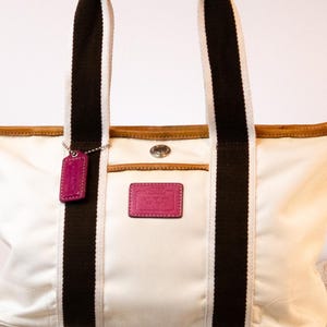 May include: Cream-colored tote bag with brown and black striped straps and a magenta leather handle. The bag features a magenta leather tag and a magenta leather patch with embossed text. The top edge is trimmed with brown leather.