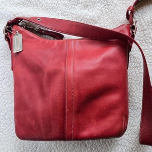 May include: A red leather shoulder bag with a long strap and silver-tone hardware. The bag has a rectangular shape with a curved top and a small tag attached to the strap. White stitching details the seams.
