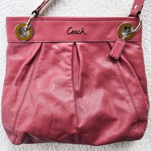 May include: A dusty rose Coach crossbody bag. The leather bag features silver-tone hardware, including rings and a small tag. The Coach logo is subtly displayed on the front. The bag has a shoulder strap and a gathered design.