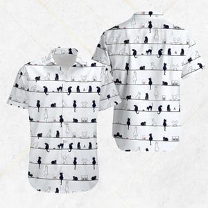 May include: White short-sleeved shirt featuring a repeating pattern of black and white cats on horizontal lines. The cats are depicted in different poses. The shirt has a classic collar and button closure.