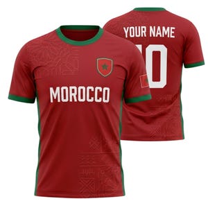 Op de afbeelding: Rood voetbalshirt met groene randen en het woord "MOROCCO" in witte blokletters. Het shirt heeft het embleem van het Marokkaanse nationale team en een kleine vlag op de mouw. De achterkant van het shirt heeft de tekst "YOUR NAME" en het nummer "10".
