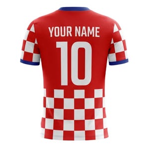 Op de afbeelding: Rood voetbalshirt met een wit geblokt patroon aan de onderkant en mouwen. Het heeft een blauwe kraag en boord. Het nummer 10 is in het wit op de achterkant gedrukt, samen met de tekst "YOUR NAME".