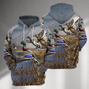 May include: A gray hoodie featuring a detailed graphic of mallard ducks in flight over a marshy landscape. The design includes ducks with green heads, brown bodies, and blue wing accents, set against a backdrop of water, reeds, and a cloudy sky.