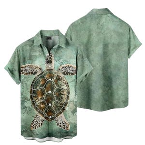 Puede incluir: Camisa de manga corta con diseño de tortuga marina. La camisa tiene una paleta de colores verde y marrón con un fondo texturizado similar a una acuarela. El caparazón y las aletas de la tortuga están detallados con patrones marrones y blancos.