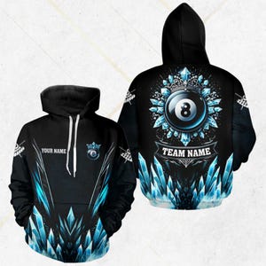 Puede incluir: Sudadera con capucha negra con temática de billar. La parte delantera presenta un área personalizable "Your Name" y un gráfico de bola de billar. La parte trasera muestra un gran diseño de bola número 8 con una pancarta "Team Name" y detalles de cristal azul.