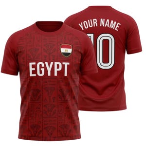 Peut inclure: Un maillot de foot de couleur bordeaux à manches courtes avec le mot "EGYPT" en lettres blanches sur le devant. Le maillot porte un écusson avec le drapeau égyptien et le numéro "10" au dos, avec le texte "YOUR NAME" au-dessus. Motif d'hiéroglyphes.