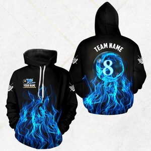 Puede incluir: Sudadera con capucha negra con un diseño de llamas azules. La parte delantera presenta un gráfico de bola de billar con el texto "YOUR NAME". La parte trasera muestra una bola ocho brillante con el texto "TEAM NAME".