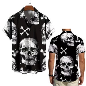 Puede incluir: Camisa negra de manga corta con un estampado de calaveras y huesos cruzados blancos. La camisa tiene un cuello clásico y cierre de botones. El diseño gótico se muestra en una percha y también lo lleva una persona.