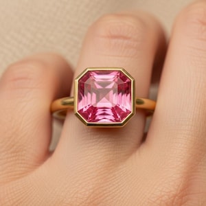 May include: A gold ring featuring a large, square-cut pink gemstone set in a smooth, polished gold bezel. The gemstone has a faceted surface that reflects light, and the band of the ring is a simple, solid gold band with a rounded profile.