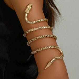 May include: Gold-toned arm cuff in the shape of a snake. The snake's head and tail are detailed, and the body wraps around the arm. The cuff is made of a textured metal, and the design is intricate and eye-catching.