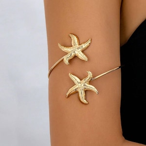 May include: Gold-colored arm cuff featuring two starfish designs. The starfish are detailed with textured surfaces and are connected by a thin, curved band that wraps around the upper arm. The jewelry is worn on a tanned arm.