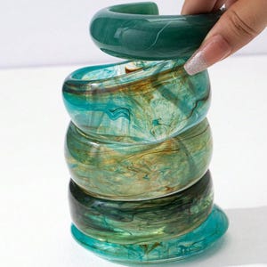 Könnte beinhalten: Ein Stapel aus fünf klobigen, marmorierten Armreifen aus Harz in Grün-, Türkis- und Brauntönen. Der oberste Armreif wird von einer Hand gehalten. Die Armreifen haben eine glänzende Oberfläche und ein Wirbelmuster.