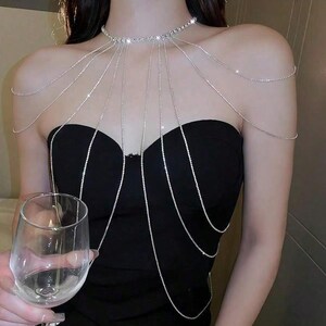 May include: A silver rhinestone body chain necklace. The necklace has a choker design with multiple strands falling over the chest and shoulders. The model wears a black strapless top. A wine glass is in the foreground.