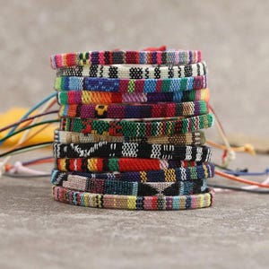 Op de afbeelding: Een stapel kleurrijke geweven vriendschapsarmbanden. Elke armband heeft een uniek patroon met een verscheidenheid aan kleuren, waaronder rood, blauw, groen, geel en zwart. De armbanden zijn ongeveer 1,3 cm breed.