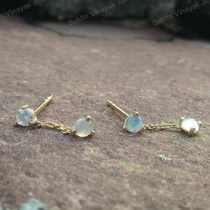 Op de afbeelding: Een paar gouden oorstekers met een kleine, ronde, iriserende edelsteen aan de pin en een bijpassende edelsteen die aan een fijne ketting hangt. De oorbellen worden gepresenteerd op een getextureerd, bruin oppervlak.