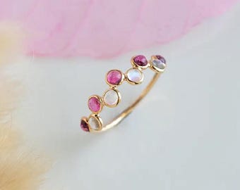 Può includere: Un delicato anello d'oro con una serie di pietre preziose rosa e bianche alternate. Le pietre preziose sono rotonde e incastonate vicine, creando un design unico ed elegante. L'anello è posto su uno sfondo morbido e sfocato.