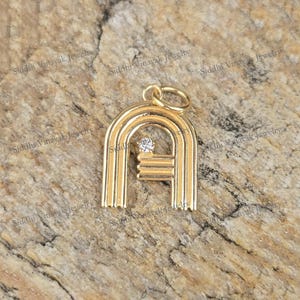 Puede incluir: Colgante dorado con forma de la letra "A", con una pequeña gema redonda y transparente. El colgante tiene una parte superior arqueada y líneas verticales, con una barra horizontal. Sobre una superficie marrón texturizada.