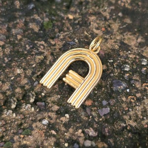 Op de afbeelding: Een goudkleurige hanger in de vorm van de letter "A". De hanger heeft een afgeronde bovenkant en een rechte onderkant met een kleine, horizontale balk. De hanger is opgehangen aan een kleine lus.