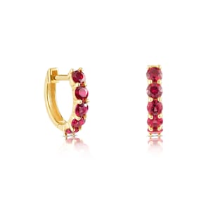 Può includere: Un paio di orecchini a cerchio dorati, ciascuno ornato da una fila di gemme rosse vibranti. Gli orecchini sono esposti su uno sfondo bianco, mostrando la loro finitura lucida e il ricco colore delle pietre.