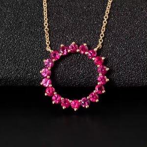 May include: A gold-colored necklace featuring a circular pendant adorned with vibrant pink gemstones. The gemstones are closely set, forming a continuous ring. The necklace chain is delicate and complements the pendant's design. The background is a dark, textured surface.