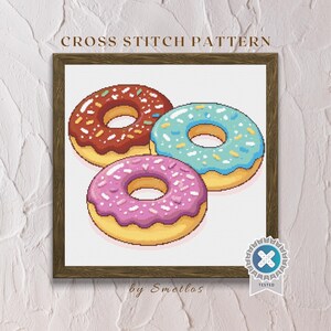 Peut inclure: Motif de point de croix encadré représentant trois beignets colorés. Les beignets ont un glaçage rose, bleu et marron avec des vermicelles. Le texte "CROSS STITCH PATTERN" est en haut.