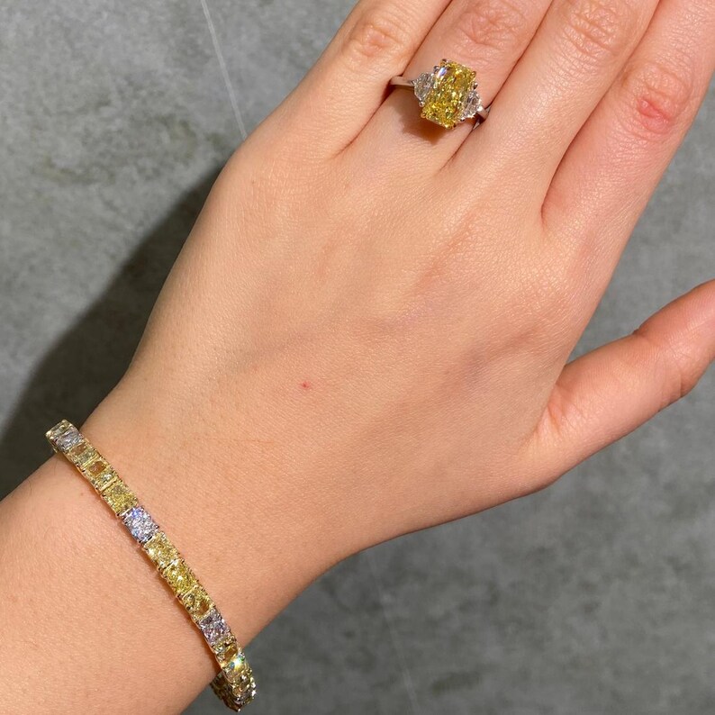 May include: A hand displays a yellow diamond ring and a matching bracelet. The ring features a large, rectangular yellow diamond with two smaller white diamonds on either side. The bracelet is a line of yellow and white diamonds.