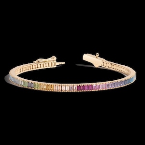 Puede incluir: Una pulsera dorada con una hilera de piedras preciosas rectangulares en colores del arcoíris. La pulsera está abierta, mostrando el mecanismo de cierre. Las piedras están dispuestas en un degradado de colores. El fondo es negro.