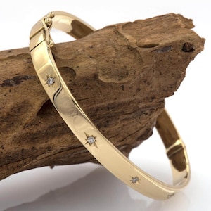 Puede incluir: Una pulsera de oro con detalles de diamantes en forma de estrella. La pulsera con bisagras se muestra sobre un trozo de madera. La pulsera tiene un acabado pulido y un cierre seguro. Es una joya clásica.