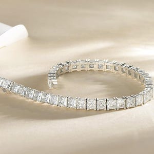 Puede incluir: Un brazalete plateado con piedras preciosas claras de corte cuadrado. El brazalete está curvado y descansa sobre una superficie beige claro. Las piedras están muy juntas, creando una línea continua de brillo.