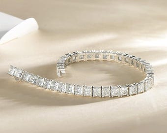 May include: A silver bracelet with square-cut, clear gemstones. The bracelet is curved and lies on a light beige surface. The gemstones are set closely together, creating a continuous line of sparkle.