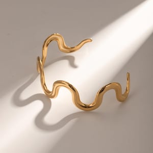 May include: A gold-colored, wavy cuff bracelet. The open-ended bracelet has a fluid, organic design with a smooth, polished finish. The bracelet's form creates a shadow on the white surface.