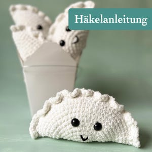 May include: A white crocheted dumpling with black eyes and a smiling mouth. Several more dumplings are in a white takeout container. The text "Häkelanleitung" is on a teal rectangle in the background.