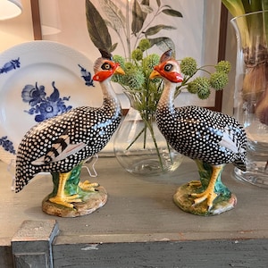 Op de afbeelding: Twee keramische parelhoen beeldjes. De vogels hebben zwart-witte gevlekte lichamen, rode en oranje gezichtsmarkeringen en gele voeten. Ze staan op groene bases. Een decoratieve schotel en bloemstuk staan op de achtergrond.