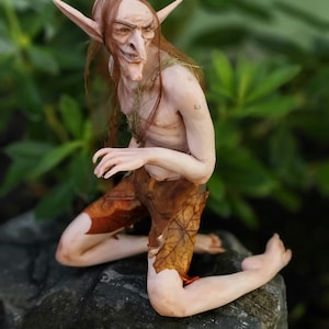 Op de afbeelding: Een fantasiewezen-figuurtje met een bleke huid, puntige oren en lang bruin haar, gekleed in een bruine broek met bladerenpatroon. Het figuurtje knielt op een donkere rots. De tekst MidDreamers Dymir staat in het wit rechtsonder afgedrukt.