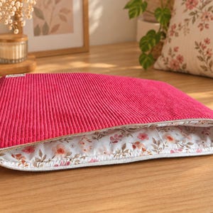 Könnte beinhalten: Eine rechteckige Reißverschlusstasche aus leuchtend pinkfarbenem Cordstoff. Das Innere ist mit einem weißen Stoff ausgekleidet, der ein zartes Blumenmuster in Rosa-, Orange- und Grüntönen aufweist. Der goldfarbene Metall-Reißverschlussanhänger ist blütenförmig.