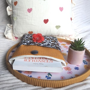 Peut inclure: Une pochette à livre avec un motif floral marron et noir, une section imitation cuir beige et une fleur rouge. La pochette est posée sur un plateau floral avec une petite plante succulente dans un pot rose et un coussin à motifs de cœurs.