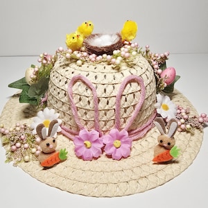 May include: A straw hat decorated for Easter with pink bunny ears, pink flowers, and small felt bunnies holding carrots. The hat also features yellow chicks, a nest with eggs, and decorative pink and white berries.