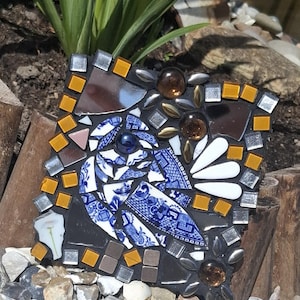 May include: A square mosaic art piece featuring a central bird figure crafted from blue and white patterned ceramic shards. The design is accented with amber glass beads, metallic silver tiles, and white petal-shaped pieces on a dark background.