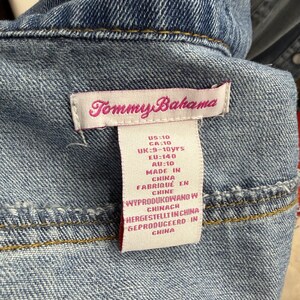 May include: A blue denim garment featuring a white fabric tag with the Tommy Bahama logo in pink text. The tag lists sizing information including US:10, CA:10, UK:9-10yrs, EU:140, and AU:10, along with manufacturing details stating it was made in China.