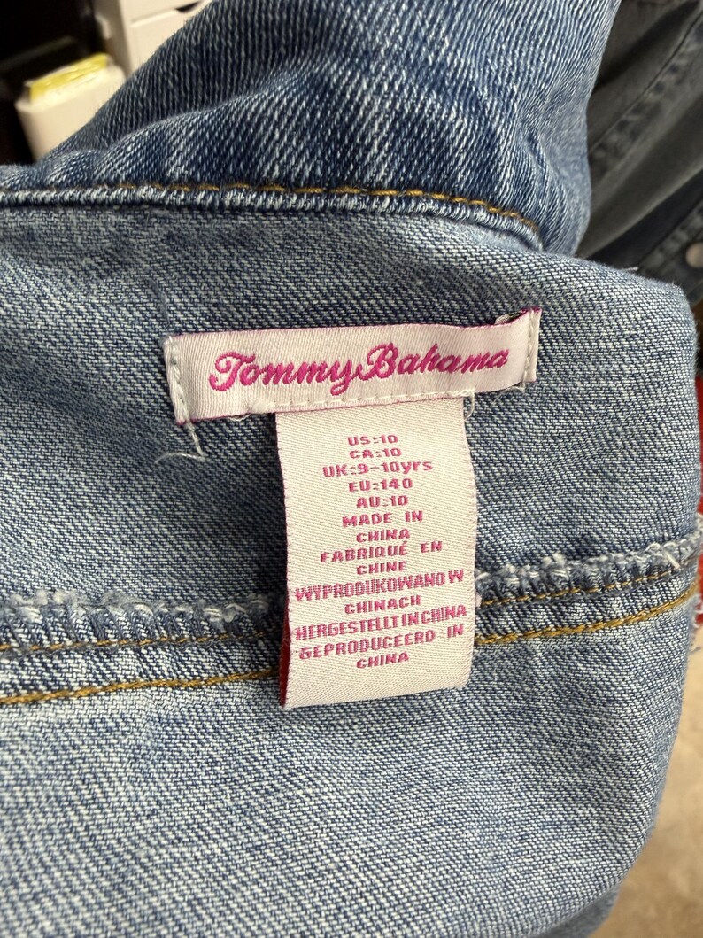 May include: A blue denim garment featuring a white fabric tag with the Tommy Bahama logo in pink text. The tag lists sizing information including US:10, CA:10, UK:9-10yrs, EU:140, and AU:10, along with manufacturing details stating it was made in China.