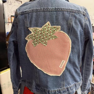 May include: A blue denim jacket featuring a large strawberry-shaped fabric appliqu&eacute; on the back. The strawberry has a red and white gingham body with a green and white floral print top, finished with a scalloped cream-colored stitched edge.