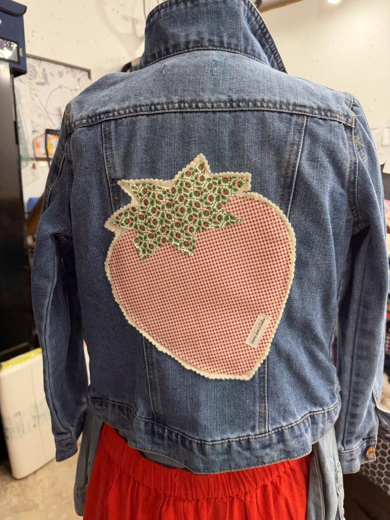May include: A blue denim jacket featuring a large strawberry-shaped fabric appliqu&eacute; on the back. The strawberry has a red and white gingham body with a green and white floral print top, finished with a scalloped cream-colored stitched edge.